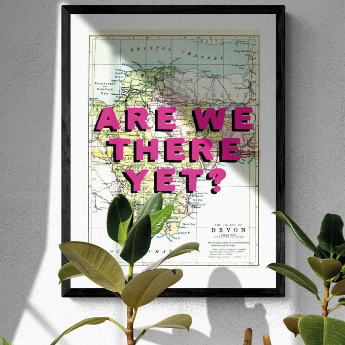 A framed Devon Map print featuring a vintage map of Devon with the playful text 'ARE WE THERE YET?' superimposed on it in pink typography.