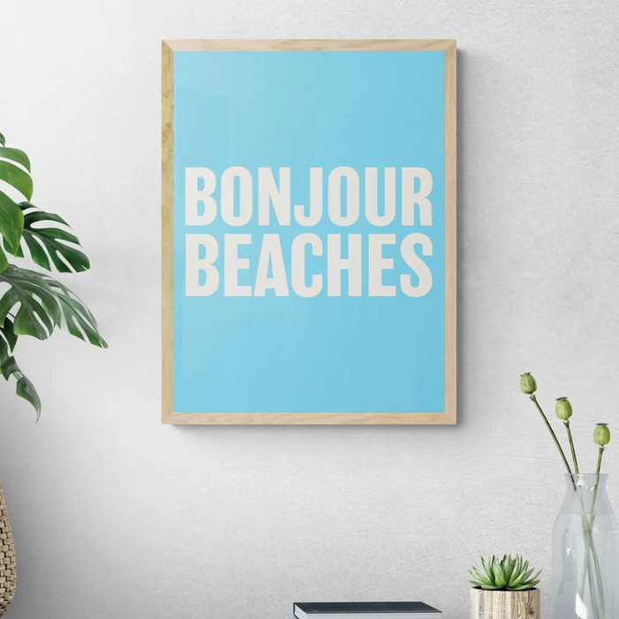 A framed word art print on the wall with a wooden frame, with the text 'BONJOUR BEACHES' in bold white letters on a blue background.