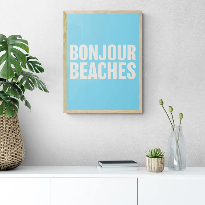 A framed word art print on the wall with a wooden frame, with the text 'BONJOUR BEACHES' in bold white letters on a blue background.
