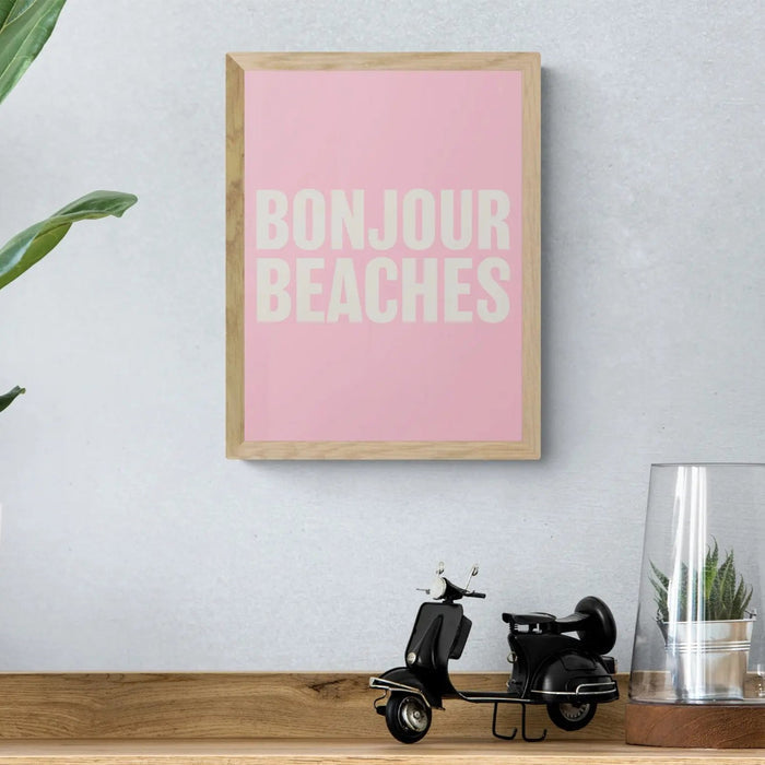 A framed word art print on the wall above a wooden shelf, with the text 'BONJOUR BEACHES' in bold, white letters on a pink background, enclosed in a black frame.