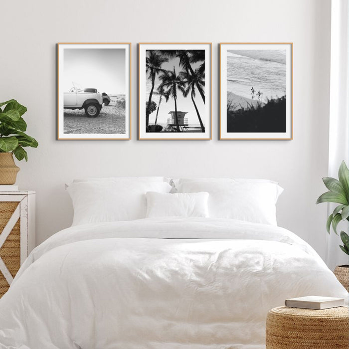 A set of three black and white coastal photography prints framed above a white bed.