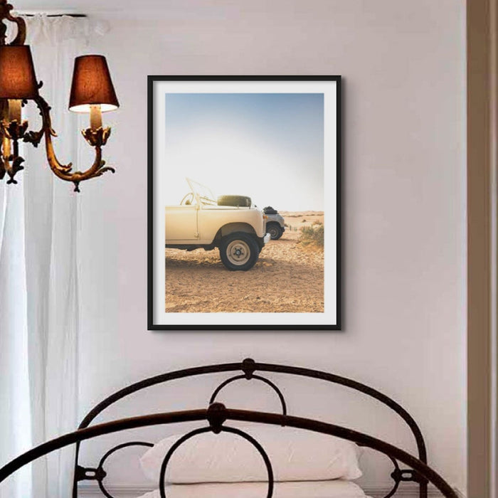 a photograph of Land Rover Defender on the beach early in the morning, in a black frame above a Victorian bed in a small bedroom with a Victorian chandelier