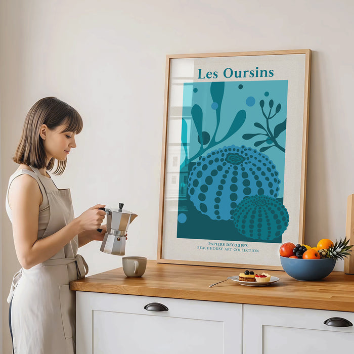 A woman making coffee in a kitchen with a framed sea urchin and seaweed art print on the wall.