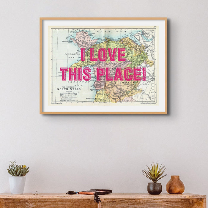 A custom map print of North Wales with a pink vintage font placeholder text saying 'I love this place!' text on a wall above a wooden shelf.