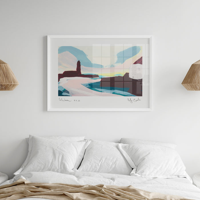 A framed abstract landscape art print of Porthleven in Cornwall hangs on a white wall above a neatly made bed with white pillows and a beige blanket. The print features soft pastel blocks of blue, pink, brown, and cream, and is flanked by two woven pendant lights.