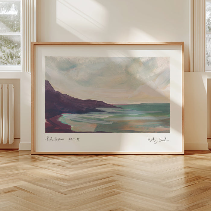 A framed landscape art print leaning against a wall in a bright room with herringbone wood floors and large windows. The painting depicts a coastal scene with muted blue-green water, a dark purple cliff, and a pale sky, set in a light wood frame.