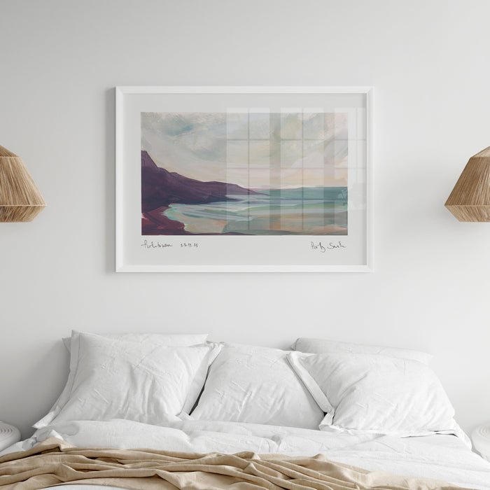 A framed landscape art print hangs on a white wall above a neatly made bed with white pillows and beige bedding. The print shows a coastal scene with soft pastel tones, and the room is styled with two woven pendant lights, a bedside lamp, and an open book.