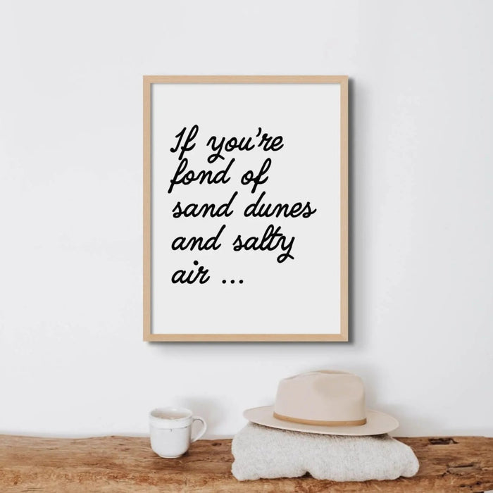A black and white typography art print with the text: 'If you're fond of sand dunes and salty air...' displayed above a bench in a beach house.