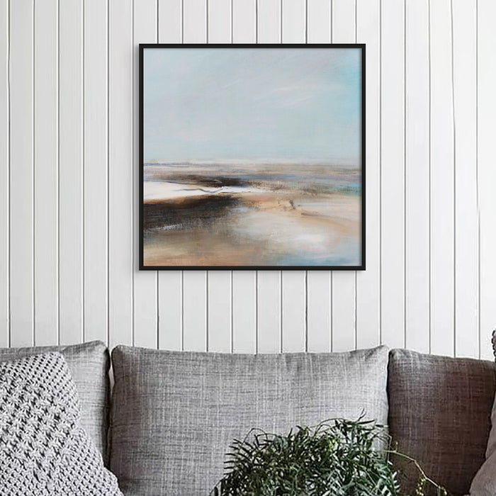 A beach painting featuring gentle coastal landscapes with soft blue and brown hues framed on wall above grey sofa.