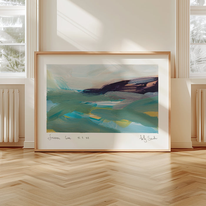 A framed abstract landscape painting leaning against a wall in a bright room with large windows, cream walls, and light wood herringbone flooring. The artwork features teal, blue, green, and dark purple brushstrokes, housed in a light wood frame with a white mat.