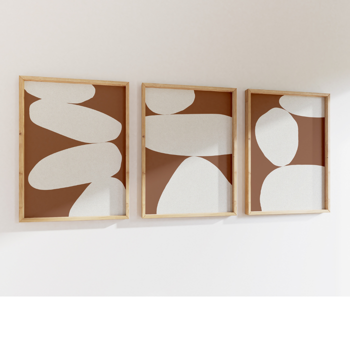Three abstract wall art pieces with white shapes on a brown background, framed in wooden frames, mounted on a white wall.