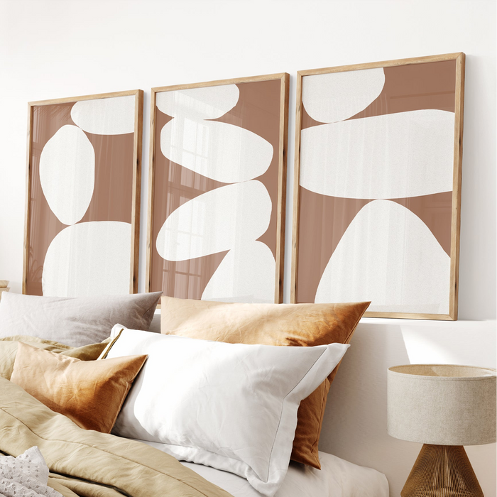 Three abstract wall art pieces with white shapes on a brown background, displayed above a bed with pillows and a lamp.