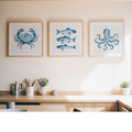 Three framed blue ocean-themed prints featuring a crab, fish, and octopus, displayed on a beige wall above a wooden counter in a modern kitchen setting.