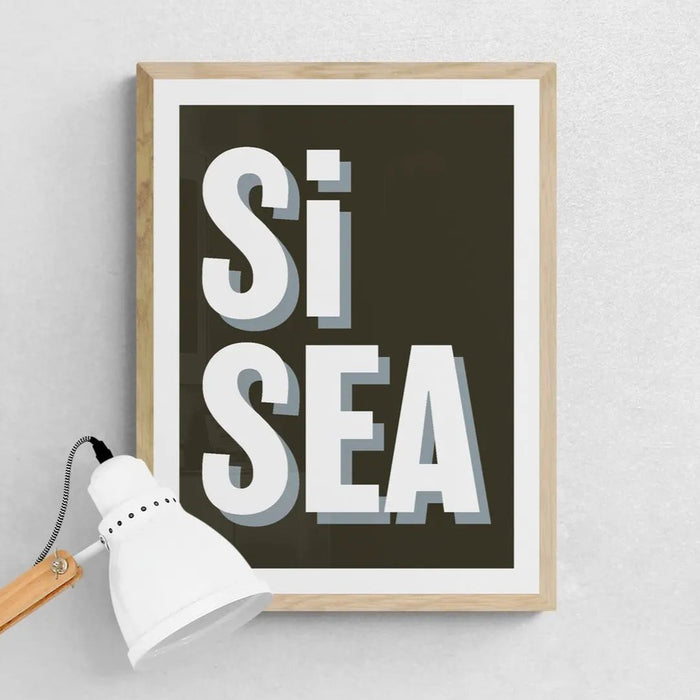 A framed black and white typography art print with the text 'Si Sea' in bold letters against a dark background.