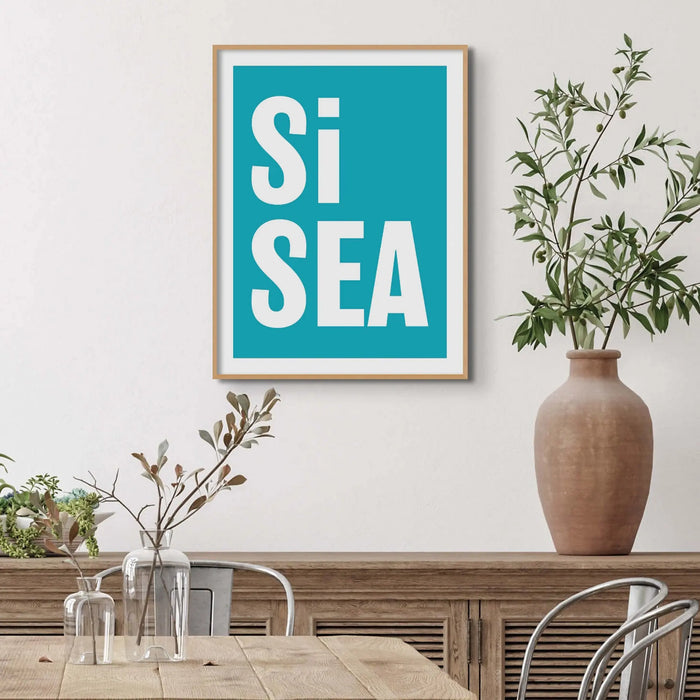 A framed typography art print with the words 'Si Sea' in large white letters on a teal background, framed in a simple natural frame.