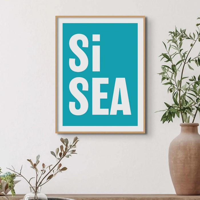 A framed typography art print with the words 'Si Sea' in large white letters on a teal background, framed in a simple natural frame.