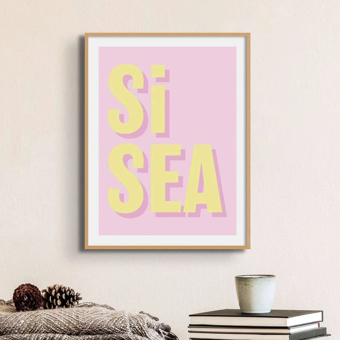 A framed typography art print with the words 'Si Sea' printed in bold yellow letters on a pink background, framed in a wood frame.
