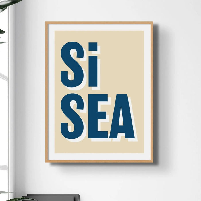 A framed coastal word art print with the text 'Si Sea' in blue letters on a beige background, presented in a simple wooden frame.