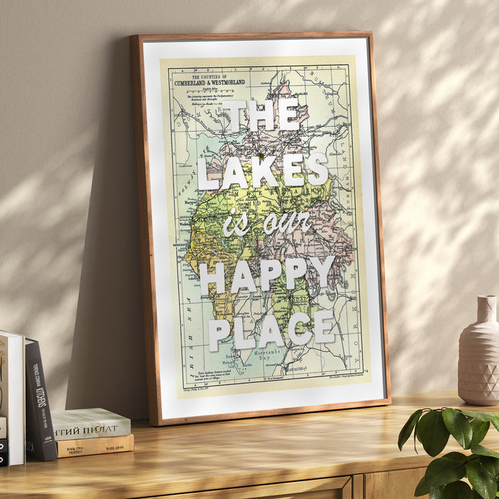 A framed map print of the Lake District on a wooden sideboard. A vintage map with the text 'The Lakes is our Happy Place' overlaid on it.