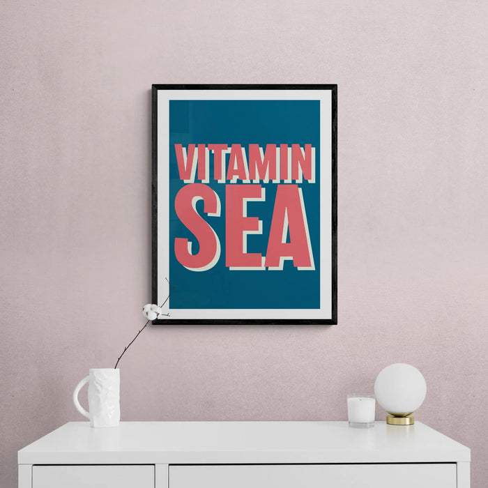 A colourful art print with the text 'VITAMIN SEA' in bold, coral-colored letters, set against a marine blue background with a black frame.