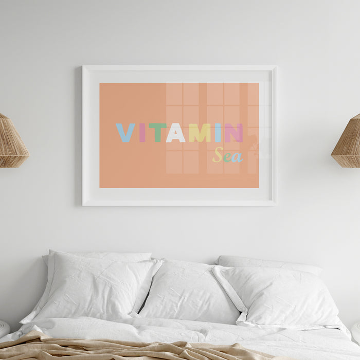 Framed art print with the text 'VITAMIN Sea' on a peach background, displayed in a white frame above a bed.